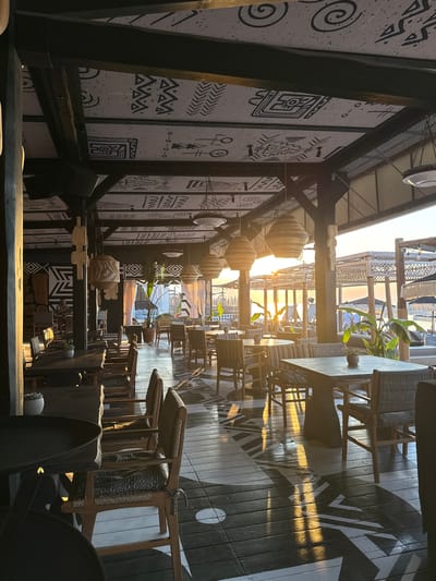 This media file captures an interior view of a restaurant or cafe, possibly with an outdoor seating area or a view towards one. The scene is dominated by wooden structures and furniture, with a striking black and white patterned wall on the left. The wall features abstract tribal-inspired motifs, including spirals, geometric shapes, and figures. Hanging from the ceiling are several large, cylindrical light fixtures or decorative elements, made of woven material.
In the background, the setting appears to be bathed in the warm, golden light of sunset or sunrise, casting long shadows across the wooden floor. The floor itself has a bold, black and white graphic design. We see rows of chairs and tables, some with woven seats and backs, and others with cushions or fabric draped over them. A few potted plants are visible, adding a touch of greenery. The architecture suggests an open-air or semi-open-air environment, with visible beams and roofing.
There are no people visible in the scene. The focus is on the objects and the overall ambiance of the space. The time of day is clearly late afternoon or early evening, indicated by the low angle of the sun and the warm light. The location is hinted at by the stylistic elements, suggesting a place with a bohemian or eclectic design aesthetic. The overall mood is peaceful and inviting, with the sunset light adding a touch of warmth and serenity. There is no visible text in the image.