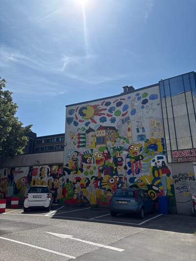 This image depicts a vibrant, colorful mural covering the side of a building in Mönchengladbach, Germany. The mural features a chaotic, cartoonish design with numerous figures, abstract shapes, and buildings. To the right of the mural, a modern building with large glass windows is visible. In front of the mural, a parking lot is present with two cars parked. A white Smart car with a license plate "MG-AK 21" is parked on the left, and a blue Ford Focus with a license plate "KK-GV 261" is parked on the right. A blue trash bin is located behind the Ford Focus. The sky is clear and blue with some thin clouds, and the sun is shining brightly, casting shadows on the ground. The presence of the Smart car with the "MG" license plate strongly suggests the location is Mönchengladbach, Germany.