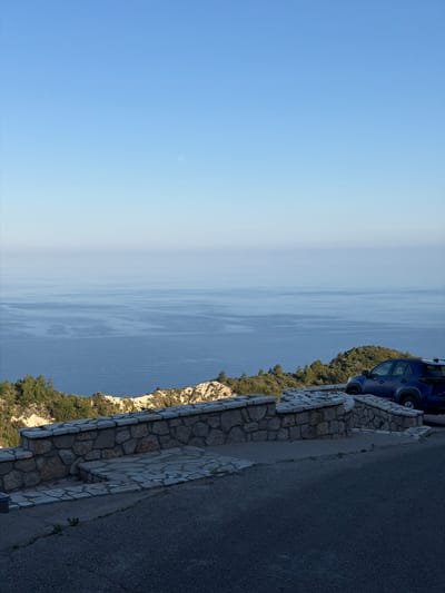 The image shows a coastal view in the Municipal Unit of Lefkada, Greece. A blue vehicle is parked along a stone wall on a roadside. The roadside runs along a cliff with greenery. Below the cliff is the sea, and above the sea is a clear blue sky.
