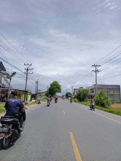 The image depicts a straight, paved road in a city, likely Payalipah, Indonesia, under a cloudy sky. The road features a yellow center line. Several individuals are riding motorcycles in the same direction. On the left side of the road, there is a building with a tiled roof and a palm tree. Utility poles with numerous wires run along both sides of the road, a common sight in Indonesian cities. On the right side, there are more buildings, some appearing to be commercial or residential, and a field of dry vegetation. The overall impression is a scene of everyday urban life in Indonesia, with a focus on transportation by motorcycle.