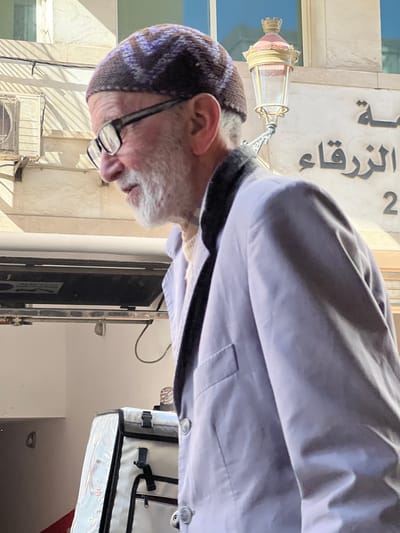 A man wearing glasses and a patterned skullcap is standing near a building with Arabic writing and the number 2 on its facade, potentially in Tanger (Tangier), Morocco. He's wearing a light-colored shirt or jacket. In the background, there is a lamp fixture attached to the building and what appears to be an air conditioning unit. Part of a delivery bag is also visible.