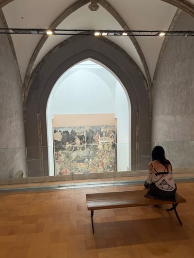 The image shows a person sitting on a wooden bench in what appears to be a museum or art gallery. The person is facing away from the camera and is wearing a black hat and a white shirt with a graphic design and ripped detailing on the back. The setting features a large, arched opening revealing a mural or painting on a distant wall. The architecture suggests an older building, possibly a church or historical site, with stone accents and high ceilings. The floor is made of light-colored wood. A glass partition separates the viewer from the art. The lighting appears to be artificial, coming from overhead fixtures and the illuminated mural. The overall mood is one of quiet contemplation and observation. The visible text on the person's shirt appears to read "VENI VIDI VIVTEO". The location is identified as Palermo, Italy, which, combined with the architecture, suggests this might be within the Norman Palace or a similar historical structure housing art.