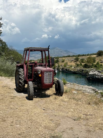 A vintage, red tractor is parked on dry, yellow grass next to a clear blue river with rocky banks. In the background, there are hills covered with sparse vegetation and a mountain range under a cloudy sky. The tractor appears to be an older model, with a metal frame and a simple canopy. The scene is serene and suggests a rural or semi-rural setting, possibly in the outskirts of Podgorica, Montenegro, known for its natural beauty and proximity to rivers and mountains. The lighting indicates it is daytime, with the sun casting shadows. There are no people visible in the image.