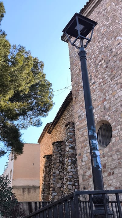 The scene captures an urban environment in Ripollet, Spain, under a clear blue sky. A prominent stone building, constructed from irregular, multi-colored fieldstones and pebbles, occupies the right and central portions of the frame. This building features a tiled roofline and three visible buttresses made of stacked stones along its side. A circular window, secured by vertical metal bars, is visible in the stone wall on the far right.

In the foreground, a dark-colored metal streetlamp with an ornate base and a modern, square LED light fixture stands slightly right of center. The lamp pole exhibits multiple light-colored, rectangular stickers adhering to its surface. To the upper left, a dense green conifer tree, displaying needles and visible cones, partially obstructs the view of the sky. In the mid-ground on the left, a building with a light orange or beige stuccoed facade is visible behind the stone structure's buttresses. A dark metal railing runs across the lower foreground, behind which small green and reddish bushes are present. No individuals or overt actions are observed.