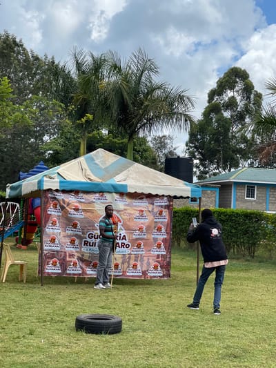 The image depicts an outdoor scene in Makuyu, Kenya, under a bright, cloudy sky during the daytime. In the foreground, a man wearing a striped shirt and grey trousers stands in front of a large banner that forms the backdrop for a white and blue striped tent. He holds a slender pole or stick. To his right, a black car tire rests on the green grass.
Further back, another man dressed in a dark hoodie and jeans stands near a simple building with a blue-tiled roof and white window frames. He also holds a vertical pole, appearing to observe the area near the tent. A large black water tank is visible adjacent to the building. The scene is surrounded by abundant greenery, including tall palm trees and dense foliage, contributing to a tropical or subtropical ambiance.
The banner prominently displays repeated text, "GUCHOKIA Rui MUKARO" and "GUCHOKIA Rui MUKARO Podcast a Kikuyu," suggesting the setting is for a podcast recording, promotion, or an event related to it. The overall atmosphere indicates an organized outdoor gathering or presentation.
