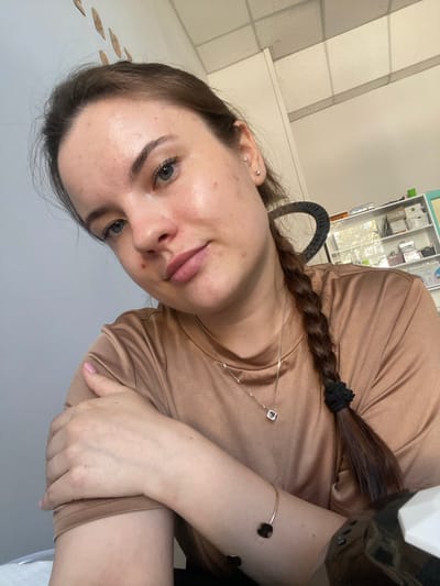 This is a portrait-oriented, close-up photograph of a young woman. She is looking directly at the camera, her head tilted to her left. The woman has fair skin, dark brown hair braided into a single plait, and light blue eyes. She is wearing a tan-colored, long-sleeved shirt. Around her neck, she wears a delicate silver chain with a small square pendant. A thin, gold-colored bracelet with dark square charms adorns her right wrist. Her fingernails are painted a light pink.
The background appears to be an indoor setting, possibly an office or salon. To the left, a wall with tiled panels is visible. In the upper right corner, a shelf unit filled with various items is partially visible, suggesting a professional environment. A black, ring-shaped object, likely part of a lamp or equipment, is also present in the frame. The lighting suggests it might be daytime, with natural light possibly contributing to the illumination. The woman's expression is neutral, with her lips slightly parted. There is no visible text in the image.