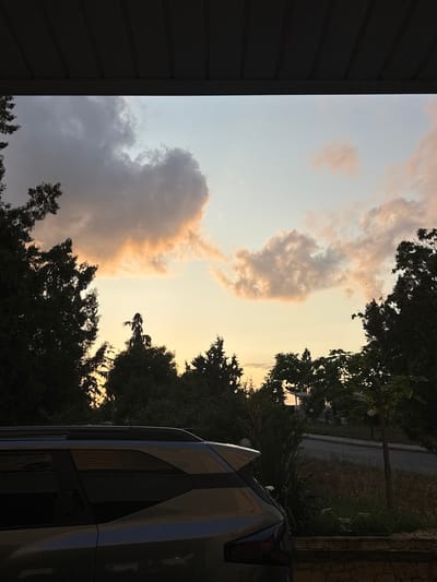 The image shows a view from under a dark, corrugated overhang, looking out at a dusky sky. Silhouetted trees, some tall and slender, others dense and bushy, frame the lower part of the sky. The sky itself transitions from a pale blue at the top to a warm, peachy orange near the horizon, illuminated by the setting sun. Scattered clouds, tinged with pink and grey, drift across the sky. In the foreground, the rear half of a silver SUV is visible, its windows dark and reflective. To the right of the car, a low stone wall runs horizontally, bordering overgrown grass and a narrow, winding road. A slender tree with a few small, light-colored spheres hangs from its branches stands near the road. The scene appears to be an outdoor residential area in Sredets, Bulgaria, possibly a driveway or a patio area, during sunset.