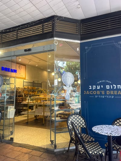 The image shows the exterior of a bakery and cafe named "Jacob's Dream" located in Ramat Gan, Israel. The facade features large glass doors and windows, offering a view into the interior where shelves are filled with bread and pastries. A sign inside the bakery displays the text "EAT WAS ALL A DREAM" with a graphic of a hot air balloon. To the right of the entrance, a dark blue wall displays the bakery's name in white and gold text, "SINCE 1986. חלום יעקב JACOB'S DREAM קונדיטוריה." Two chairs with intricate woven patterns and a small round table with a speckled surface are positioned outside the entrance, suggesting an outdoor seating area. The street in front of the bakery is paved with red bricks and concrete. The overall lighting suggests it is daytime.