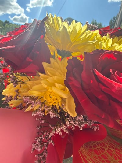 The image depicts a close-up view of a bouquet of flowers with red roses and yellow chrysanthemums, possibly taken in an outdoor setting due to the presence of a bright sky and glimpses of trees in the background. The flowers are arranged with smaller purple filler flowers and wrapped in red decorative material, possibly paper or cellophane. The close proximity of the camera to the bouquet allows for a detailed examination of the flowers' textures and arrangements, but it provides limited information about the broader surroundings or specific actions taking place in the image. There is no definitive evidence in the image to confirm that the location is Dmitrov, Russia.
