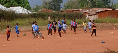 A group of children play soccer on a dirt field in Ngalula, Democratic Republic of the Congo. Several children wear blue and red soccer jerseys. One boy kicks a green soccer ball. In the background are small buildings with corrugated metal roofs and trees. A girl in a white dress stands in the middle of the background near the building to the left. A blue water bottle is on the ground near the right of the image.