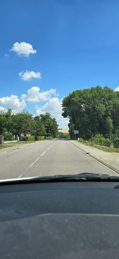 A daytime, eye-level, forward-facing outdoor shot shows a paved road leading into the distance. The road has a dashed white line down the center and a white arrow indicating a lane. The road is bordered by a sidewalk on the right and grass and trees on the left. In the distance, fields can be seen. The sky is blue with scattered white clouds. A signpost with directional arrows is visible on the right side of the road. The lower portion of the image shows the dashboard of a vehicle, including the windshield wipers. The image appears to have been taken from inside a car driving on the road. The signpost indicates the towns of "Villingen" and "Schaffhausen". Given the context of Villingen and Schaffhausen, this location is likely in Germany or Switzerland, not France.