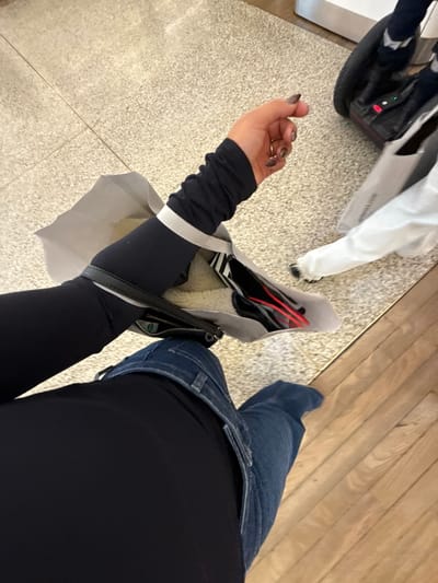 A high-angle photograph captures a scene within an indoor commercial establishment in Saint-Laurent-du-Var, France. The primary subject, seen from the waist up to the forearm, is wearing a dark long-sleeved garment and blue denim jeans. The subject's right forearm, featuring manicured nails with dark polish and a gold-colored ring on the ring finger, holds a light gray paper shopping bag with white handles. Inside the bag, dark objects with red elements and white packing material are visible. A small black zippered pouch is positioned on the subject's left thigh, partially obscured. The floor in the foreground is composed of light-colored, speckled tile, transitioning to light brown wooden floorboards in the mid-ground, separated by a thin metallic strip. In the background, to the right, are the lower legs of a second individual wearing white trousers and dark shoes, positioned near a black self-balancing scooter (possibly a Segway or hoverboard) which displays illuminated red and green indicators. A second white shopping bag is partially visible near the second individual's legs.