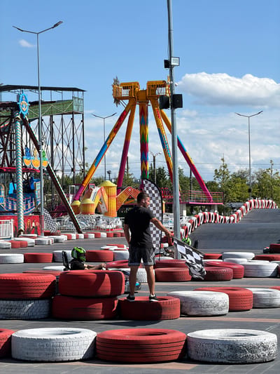 In Tohatin, Moldova, an amusement park features a go-kart track bordered by stacked tires painted red and white. A man stands on a tire, holding a checkered flag, facing a go-kart. Another go-kart, driven by a person wearing a green helmet, is positioned on the track. In the background, amusement rides with colorful structures are visible under a bright blue sky with scattered clouds.