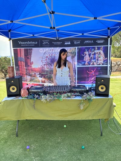 A vertical, outdoor shot shows a DJ performing in Pueblo Nuevo de Jasso, Mexico. The DJ is a young woman wearing a white-striped shirt. She is standing behind a Pioneer DJ setup on a table covered with a green tablecloth, which is decorated with flowers. The DJ is adjusting the controls on the mixer. Behind the DJ setup is a large banner with the logos of various brands, including VidantaWorld, Cirque du Soleil, and Jungala. The banner features images of the attractions offered by these brands. The DJ setup is flanked by two KRK speakers. The DJ is under a blue tent. The ground is covered in artificial turf. Several golf balls can be seen on the turf.
