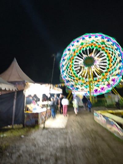 The image displays a nighttime scene at a fairground or market in An Khe Ward, Vietnam. A large, rotating amusement ride, consistent with a Ferris wheel, occupies the right side of the frame. This structure is heavily illuminated with multicolored lights in patterns of green, blue, purple, yellow, and white, exhibiting significant motion blur. To the left, a light-colored, conical-roof tent is partially visible. Adjacent to it stands a smaller, open-fronted stall with dark side coverings and bright internal lighting. Within or near this illuminated stall, approximately 5-7 individuals are present, their features indistinct due to blur. A dirt or gravel pathway extends through the foreground and mid-ground. Along this path, 3-4 additional individuals are observed walking away from the camera, towards the right, their figures heavily blurred. Faint string lights or banners are visible strung above the stall area. The sky is dark, indicating a nocturnal environment.