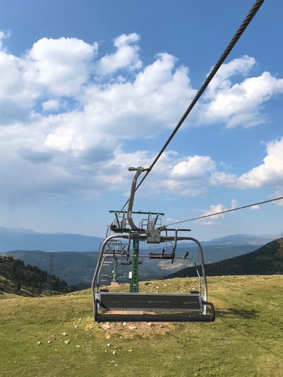This is a medium shot of a ski lift chair in the foreground, looking out over a mountainous landscape under a partly cloudy sky. The ski lift chair is empty and appears to be suspended from a thick cable that runs diagonally across the frame from the top left to the bottom right. The chair is made of metal and has a dark seat and backrest. In the background, rolling hills covered in green grass and sparse trees descend into a valley. Further in the distance, more layered mountain ranges are visible, their peaks receding into a hazy blue. The sky is a vibrant blue with scattered white, fluffy clouds. The overall scene suggests a bright, clear day, likely during the summer or autumn months, as there is no snow visible. The location is identified as Bagà, Spain.