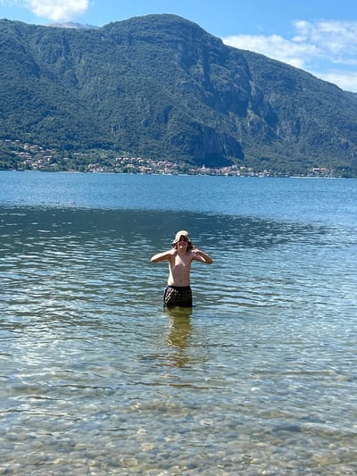 The image displays a male youth standing chest-deep in the clear, rippled waters of Lake Como, specifically in the area of Abbadia Lariana, Italy. The lakebed, composed of numerous rounded stones, is visible through the transparent water. The individual, shirtless and wearing dark, patterned swim trunks and a light-colored bucket hat, faces forward with a visible smile. His arms are bent at the elbows, and both thumbs are pointed downwards. In the background, across the expanse of the lake, a series of densely forested mountains rise, with a village or small town composed of light-colored buildings with red roofs nestled along the distant shoreline at their base. The sky above is bright blue with scattered white clouds.