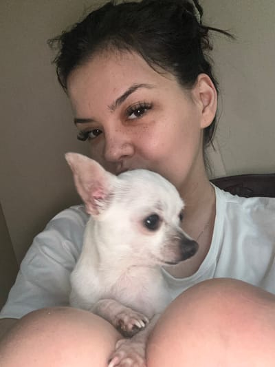 The image captures a close-up, intimate moment between a person and a small dog, presented as if the camera were held sideways or the subjects were lying down. A person with dark, slightly messy hair, visible ear, prominent dark eyelashes, and subtle freckles on their face is seen from the neck up, wearing a white t-shirt and a delicate silver chain necklace. They are looking directly towards the camera with a soft, affectionate expression.
Snuggled against the person's face and upper body is a small white dog, likely a chihuahua or similar breed. The dog has mostly white fur, dark eyes, and pointed ears. Its head is turned slightly, also looking towards the camera. A notable detail is a reddish, possibly irritated, patch visible on the inside surface of one of the dog's ears.
The scene takes place indoors, suggested by the plain, light-colored background and soft, diffused lighting. The person appears to be lying on a soft, white surface, possibly a bed or couch, which contributes to the relaxed and comfortable atmosphere. The overall mood is one of tender companionship and peace. There is no visible text in the image.