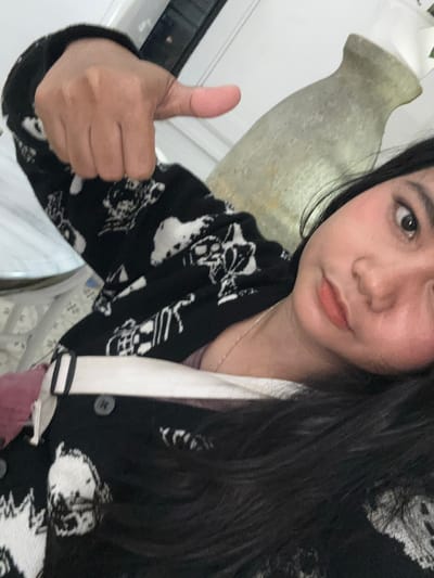 A young woman with long, dark hair and makeup is lying down and taking a selfie. She is wearing a black and white patterned cardigan over a white top. Her left hand is raised, giving a thumbs-up gesture. In the background, a light-colored, elongated vase or urn is visible. The scene appears to be indoors, with a white wall and furniture elements suggesting a domestic or possibly a cafe setting in West Jakarta, Indonesia.