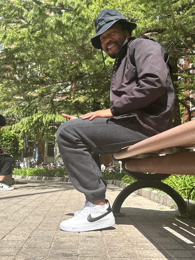 A man sits on a park bench. He wears a black bucket hat and a dark jacket. He has gray sweatpants and white sneakers with a black Nike swoosh. He is smiling and holding a phone. The bench is metal and wood and is located in a park with trees and grass. The sidewalk is paved with stone. There is no indication of the location being Daxing District, China.