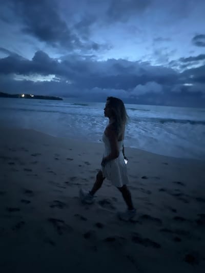 The image depicts a lone woman standing on a sandy beach at dusk or during a storm. She is wearing a light-colored dress that appears to be white or off-white. Her hair is light-colored and she is facing towards the ocean. The waves are gently rolling onto the shore, and the sand is imprinted with numerous footprints.
The sky is filled with dark, dramatic clouds, suggesting either an impending storm or the aftermath of one. The lighting is dim, contributing to a moody and atmospheric feel. In the distance, along the coastline, a few scattered lights are visible, possibly indicating a town or resort. The overall scene is one of solitude and a connection with nature's raw elements. The location is identified as Choeng Thale, Thailand.