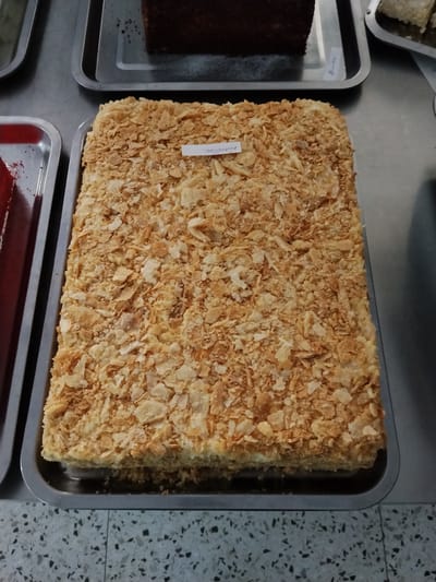 A rectangular Napoleon cake, covered in flaky pastry crumbs, rests on a metal tray. A white paper label with black text, likely a product name or description, is placed on top of the cake. The cake is presented in what appears to be a commercial setting, possibly a bakery or a food market, with other food items and trays visible in the background. The environment is likely indoors, as suggested by the artificial lighting and the tiled floor. There are no people visible in the image. The image captures the cake from a slightly elevated, frontal perspective.
