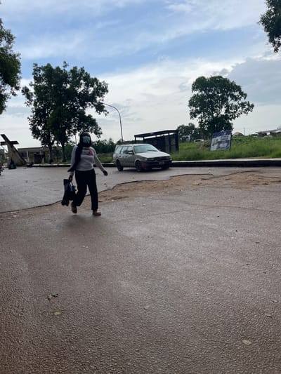 A person wearing a light-colored long-sleeved shirt, black pants, and a face mask is shown in motion, seemingly practicing dance or freestyle moves on a wet asphalt road. They are holding a dark-colored bag. The scene is set in Rafin Par, Nigeria, under a cloudy sky suggesting it might be overcast or after rain. Mature trees with dense foliage line the background, with one large tree prominently featured. A white car is parked near a damaged roadside curb. A street lamp is visible, and a sign with text that includes "STITCH" and an image is posted behind a tree. The overall atmosphere appears casual and the person's pose suggests an active, perhaps artistic or recreational, moment.
