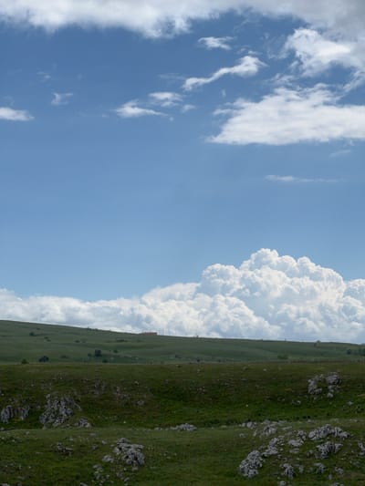 The image shows a view in Pišče, Montenegro. A grassy, rocky hillside occupies the right side of the frame. Above the hillside, a large cumulonimbus cloud formation rises into the sky. The left side of the image shows a blue sky with scattered cirrus clouds.
