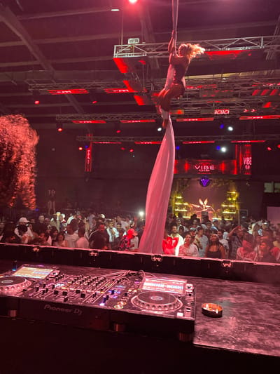 The image depicts an aerial performer suspended by a rope against a backdrop of a crowded nightclub. The nightclub features red and pink lighting, a DJ booth with Pioneer DJ equipment in the foreground, and a large crowd of people dancing and observing the performance. A sign above the crowd reads "VIBE CLUB." The venue is located in Sharm El Sheikh, Egypt. The aerialist, a blonde woman in a dark leotard, is performing with white fabric. The crowd appears to be diverse, with many individuals holding up cell phones to record the event. The overall atmosphere is lively and energetic, consistent with a nightclub setting.