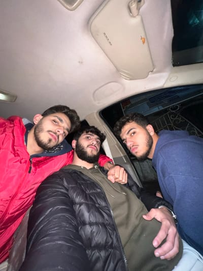 Three young men pose inside a vehicle, likely a van or car. The perspective is from the back seat. The man closest to the camera wears a red jacket. The man in the middle has a black puffer jacket over a green hooded sweatshirt, and his fist is clenched. The third man, partially obscured, wears a blue sweatshirt. The vehicle's interior is visible: a beige sun visor and ceiling, and a glimpse of the outside through the window.