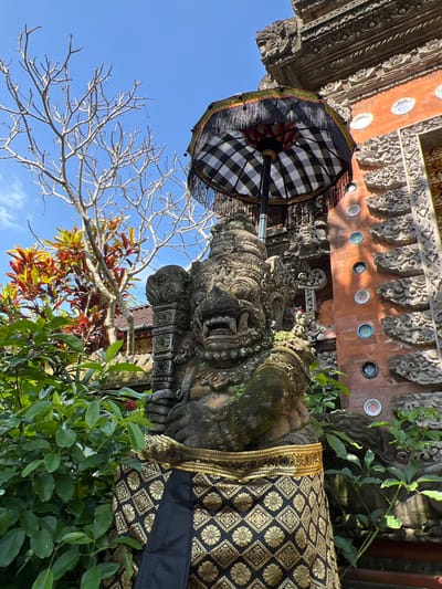 A ground-level shot in Petulu, Indonesia, angled upward toward a stone statue adorned with a black and gold patterned cloth and a black and white checkered umbrella. The statue has detailed facial features and mossy texture. A bare tree branch and lush green foliage frame the statue. Behind the statue is an ornately carved structure with a row of circular embellishments on a red background. The sky is visible in the upper left corner and is bright blue.