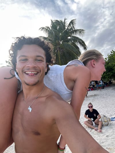 The image captures three individuals on a beach, likely in Sainte-Anne, France, based on the tropical vegetation visible, which suggests a coastal or island location in the French West Indies. The primary subject is a man in the foreground taking a selfie. He has curly brown hair, tanned skin, and is smiling, showing his teeth. He's wearing a silver necklace with an island pendant. Behind him and to the left is a woman in a white tank top, her head thrown back in laughter. A tattoo is visible on her upper arm. In the background, another woman sits on the beach, wearing sunglasses and a black t-shirt, looking at her phone. There is a beach bag near her. The environment includes palm trees, white sand, and other people further away from the camera. The sky is partly cloudy.