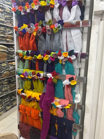 This is a medium-shot, eye-level, indoor photograph of a retail display of colorful fabric scarves, possibly in a market stall or shop, located in the Ubud District, Indonesia. The scarves are neatly folded and hung on tiered metal racks against a white background, with shelves of more fabric goods visible in the blurred background to the left. The foreground features a collection of brightly colored scarves, many adorned with decorative fabric flowers that have pearl centers and gemstone accents. The scarves are predominantly in shades of orange, yellow, purple, green, and grey, with some also featuring white and pink. The lighting is even and bright, illuminating the vibrant hues of the products. There are no people visible in the image. A security tag with the price "Rp. 10,000" is attached to one of the scarves. The overall impression is of a well-organized and visually appealing display of textile merchandise.
