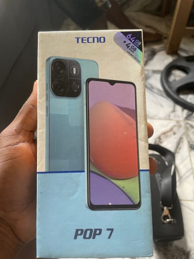 A light blue cardboard box for a TECNO POP 7 smartphone is being held by a person's left hand. The box features a picture of the phone on the front, showing its light blue back with a dual-camera setup and a purple and orange-toned screen. A sticker in the upper right corner of the box indicates "64GB ROM + 4GB RAM extended 2GB RAM". The background is out of focus but appears to be an indoor setting with tiled flooring and furniture. The person's hand is dark-skinned and partially visible on the left side of the image. The scene is likely set in Keffi, Nigeria, given the context of the product.