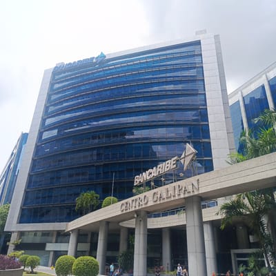 The image shows the Bancaribe building, located in the city of Caracas, Venezuela. The modern skyscraper features a distinctive blue glass facade and a prominent sign that reads "BANCARIBE". Below the main building, an entrance area is visible, with the sign "CENTRO GALIPAN" displayed over the walkway. Several individuals are present in front of the building, some are sitting and others are standing. The area is landscaped with trees and bushes, contributing to an urban park-like setting. The sky is overcast, suggesting a cloudy day. The architectural style of the building suggests a contemporary commercial or financial institution.