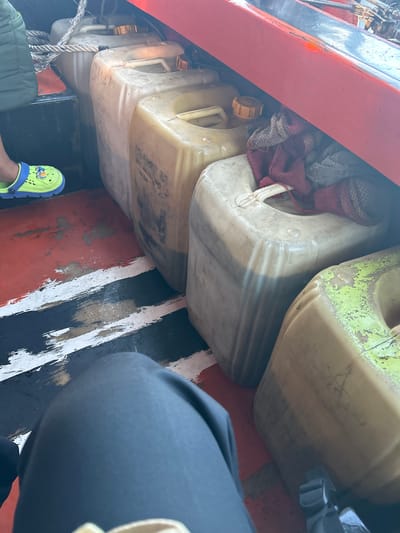 In an unknown location in Indonesia, multiple plastic jerrycans, some filled with a dark liquid, are stored in the back of a vehicle, possibly a boat. A person's legs are visible in the foreground, one wearing a lime green clog with cartoon characters. Ropes are securing the jerrycans. The floor of the vehicle has a painted pattern of red, white, and black stripes. A red railing is visible above the jerrycans.