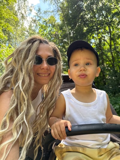 In Rusanovo, Russia, a woman with long, blonde dreadlocks and sunglasses is captured in a selfie with a young child. The woman is wearing a white tank top, and the child is wearing a white tank top and a black baseball cap. They are both outdoors, with lush green trees and a blue sky visible in the background. The child appears to be sitting in a stroller, with the woman partially obscured by the stroller's handle. The overall scene suggests a casual outing on a sunny day.