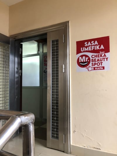 The image shows a doorway with a metal door and a sign on the wall next to it. The doorway is located on a landing of a staircase, and the door is open to reveal a tiled hallway. The sign is a red rectangle with white text that reads "SASA UMEFIKA kwa Mr. CHEKA BEAUTY SPOT HAPA."  The sign is a sign for a beauty salon and is located at the coordinates -3.372, 36.689 in Arusha, Tanzania. The landing has a metal railing and the wall is beige and cracked in places.  There are no people in the scene.