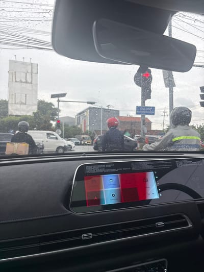 A car interior view shows a digital dashboard display featuring red and blue square icons. Through the rain-streaked windshield, a street scene includes a red traffic light, utility poles, and various pedestrians and motorcyclists, some wearing helmets. A white van is also visible. The rearview mirror is positioned above, reflecting the street scene.