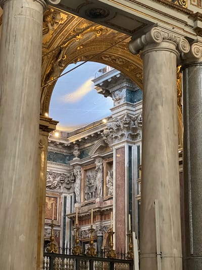 This image captures the opulent interior architecture of the Basilica Papale di Santa Maria Maggiore in Rome, Italy. The foreground is framed by two substantial marble columns: a light beige-grey column on the left and a similar column with an Ionic capital on the right, partially obscuring a darker column behind it. Centrally, a large gilded archway, richly adorned with intricate gold relief work and sculpted figures, spans between the columns; one figure appears to be dynamically suspended from the upper left interior curve of the arch. Through this arch, a further section of the basilica is visible, featuring a vaulted ceiling covered by a light-colored protective netting, illuminated by several artificial spotlights. The background architecture displays walls paneled with polished dark green and reddish marble, contrasting with white marble elements and elaborate white stucco or carved stone decorations. Numerous classical sculptures are integrated into these walls, including winged putti, and larger figures standing within niches. At the bottom, a dark metal balustrade supports multiple ornate golden candelabras, each holding a white candle. No individuals are present in the scene.