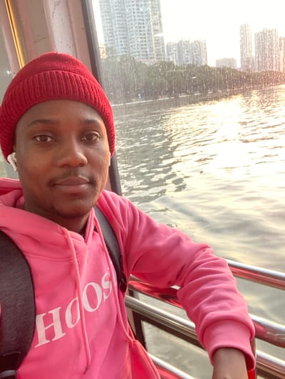The image shows a close-up portrait of a young adult, likely male, aboard what appears to be a boat or ferry. He has dark skin, is wearing a bright red knitted beanie, and a vibrant pink hooded sweatshirt. White wireless earbuds are visible in his ears. He looks directly at the camera with a neutral expression. Dark backpack straps are visible over his shoulders.

The background features a wide body of water, possibly a river or bay, brilliantly reflecting golden, warm light, suggesting either sunrise or sunset. The water's surface is covered in ripples. Beyond the water, a dense and modern cityscape rises, characterized by numerous tall buildings forming a prominent skyline. Some greenery and what appears to be a road are visible along the distant bank. Silver metal railings, indicative of a boat's structure, are present to the right of the person.

The overall atmosphere is illuminated by the soft, glowing light of dawn or dusk. Part of a white logo, spelling "SOOH", is visible on the front of his pink hoodie. This scene is set in Guang Zhou Shi, China, depicting a moment on the water with the city as a backdrop.