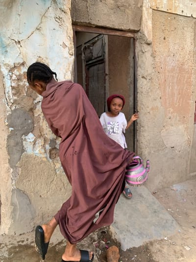 This image depicts an outdoor scene in Gwagwarwa, Nigeria, featuring two females positioned against weathered building exteriors.
In the foreground, an adult or older female is captured from the back, in motion, appearing to walk or pivot towards the left. She wears a long, loose-fitting, reddish-brown garment that extends to her ankles. Her hair is dark and styled in braids, and she wears small gold hoop earrings. On her feet are black slide sandals. Her right hand holds a silver metallic teapot adorned with prominent pink horizontal stripes, a pink handle, and a pink spout.
In the background, a young female child stands within a recessed doorway, looking directly at the camera with a neutral expression. She wears a white short-sleeved t-shirt featuring a stylized graphic design on the chest. A dark red head covering conceals her hair. Her right arm is slightly raised, with her hand near the door frame. Her feet, visible beneath the doorway, wear sandals with pink straps over striped socks.
The immediate environment consists of dilapidated building walls. The wall to the left of the foreground subject exhibits flaking plaster, revealing underlying light blue and brown layers. The wall to the right, forming part of the doorway, is a textured, light brown, with faded vertical white chalk-like markings. The ground is an uneven mixture of dirt and concrete, with scattered small debris including reddish-brown fragments. The doorway itself is framed by rough concrete or stone and contains a dark, possibly wooden, recessed door. The scene is illuminated by natural daylight.