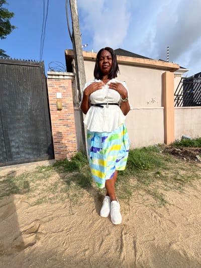 A single adult female is depicted standing outdoors on a sandy patch of ground in Iranla, Nigeria. She faces the viewer directly, with her eyes partially closed and her mouth slightly downturned. She wears a white, textured short-sleeved blouse, a thin black belt, a long silver-colored necklace with a dark pendant, a multi-colored striped skirt (blue, light green, yellow, white), and white athletic shoes. Her hands are positioned over her upper chest/abdomen.
The background consists of a residential street scene. To the immediate left is a tall, dark metal gate with a diamond plate pattern, adjacent to a brick pillar topped with coiled barbed wire. A light-colored electrical box and a brass-colored light fixture are mounted on this pillar. A concrete utility pole with overhead electrical wires stands centrally in the mid-ground. Behind the subject and to the right are various residential buildings with light-colored, possibly beige, exterior walls and dark roofs. One building's roofline shows a striped black and white pole. The ground is dry earth with sparse green grass patches. The sky is partly cloudy with areas of clear blue visible.