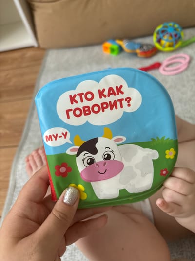 A close-up, overhead shot captures a partially seen baby being held by a person, who is presenting a children's book. The book, with a blue sky and green grassy background, features a cartoon cow. Text on the book reads "КТО КАК ГОВОРИТ?" and "МУ-У". The baby's legs and feet are visible at the bottom of the frame. The person's hand, with glittery nail polish, is holding the book. In the blurred background, other baby toys, including a colorful rattle and pink rings, are scattered on a light gray mat. The scene appears to be indoors, possibly in a nursery or playroom, given the presence of baby items. The lighting suggests it's daytime. The overall impression is one of playful interaction between the baby and the adult, with the book serving as the focal point of the activity.