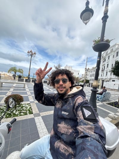 A male individual is depicted in a medium shot, smiling directly at the camera and making a "peace sign" with his right hand. He possesses dark, curly hair and a beard, and wears round sunglasses. His attire consists of a hooded jacket featuring a distressed camouflage or abstract pattern in shades of navy blue, light blue, and tan, with a visible light-colored hood lining and a black patch with a white mountain motif on the right sleeve. He is also wearing light blue ripped jeans and white shoes. The subject is seated on a light grey/white scooter, with its handlebars and rearview mirror visible in the lower left quadrant.
The scene takes place outdoors in **Place du 9 Avril 1947 (Grand Socco) in Tangier, Morocco**. The plaza floor is paved with a distinctive black and white checkered tile pattern, interrupted by planted garden beds containing white flowers and green foliage. In the mid-ground, an ornate dark streetlamp with gold accents and an attached flower planter is positioned to the right. A second similar streetlamp is visible further back on the left. A large, dark cannon is situated in the mid-background, left of the central subject. The background features several white, multi-story buildings exhibiting traditional Moroccan architecture. A prominent white historic building with multiple arched windows is visible in the far background. The sky above is partially cloudy, revealing patches of blue. One male is observed seated on a distinctive, colorful patterned bench in the mid-ground to the right. Several other indistinct figures are present in the distant background.
