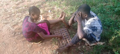 Two young individuals are seated on the ground outdoors in Ngalula, Democratic Republic of the Congo, playing a game of Mancala. The girl is wearing a pink dress with a black, patterned overlay, while the boy is wearing a t-shirt and pants. They are both barefoot. The Mancala board, made of wood with marble pieces, is placed between them on the ground, which consists of dirt and some patches of grass. The boy has his hand to his head, appearing deep in thought.