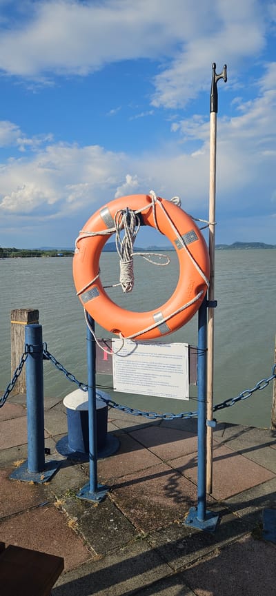 A life preserver ring hangs on a wooden pole next to a blue chain fence on a pier. The life preserver is orange with reflective silver markings and a coiled white rope. A sign is attached to the pole below the life preserver. The pier is paved with reddish-brown and gray interlocking pavers. A dark blue mooring bollard is visible behind the fence. The water is calm and reflects the sky, which is blue with scattered white clouds. In the distance, a green, hilly shoreline is visible with trees. The scene is located in Balatongyörök, Hungary.
