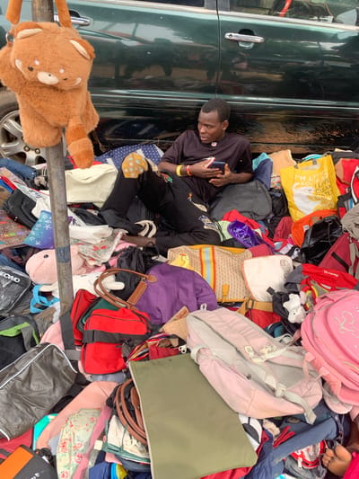 A young man is lying down amidst a large pile of bags, backpacks, and luggage. He is wearing a black t-shirt and dark pants, and is holding a smartphone in his right hand. He appears to be looking at the phone. The bags are of various colors and sizes, including pink, yellow, purple, red, green, and black. Some are brightly colored, while others are more subdued. There are also some stuffed animals visible in the pile, including a teddy bear and a pink whale. The scene appears to be outdoors, possibly a street or a market, given the presence of a dark green vehicle on the left side of the frame with a chrome door handle and a car wheel visible at the bottom. The ground is covered in bags and the overall impression is one of abundance or perhaps a chaotic sale or storage situation. The lighting suggests it is daytime. There is text on a yellow bag that reads "I'M LITT & CHILD FRIENDLY".