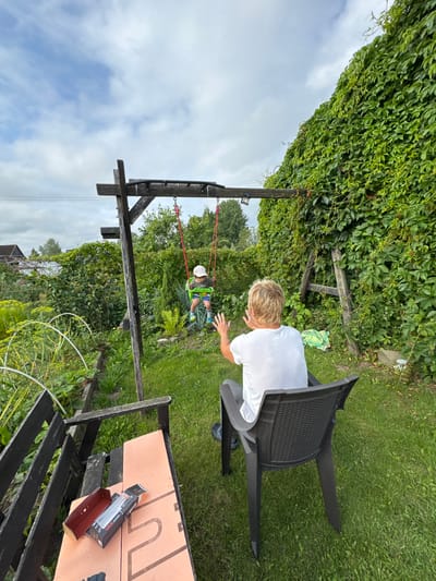 In the backyard of a property in Sitno, Russia, two individuals are present. In the background, a child sits on a swing, propelled by an unseen force, wearing a light-colored cap and blue shorts. In the foreground, an adult with blonde hair, dressed in a white t-shirt, sits in a dark plastic chair, facing away from the camera and towards the child on the swing, with their hands raised. The yard features lush green grass, a wooden swing set structure, and a dense wall of green ivy covering a wooden fence or structure. Partially visible in the lower left is a wooden bench with a box and packaging on it. The sky above is partly cloudy.