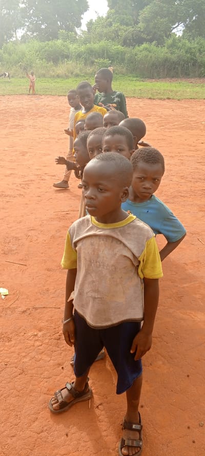 A line of about ten African children stands on a red dirt surface in Ngalula, Democratic Republic of the Congo. They are of varying heights and wear casual clothes. The child in the front wears a beige t-shirt with yellow sleeves and dark blue shorts, with sandals. Some children are barefoot and wearing tattered clothes. In the background, a grassy area with trees can be seen. In the distance, a goat can be seen grazing, and there are two other people. The scene appears to be outdoors and somewhat impoverished.